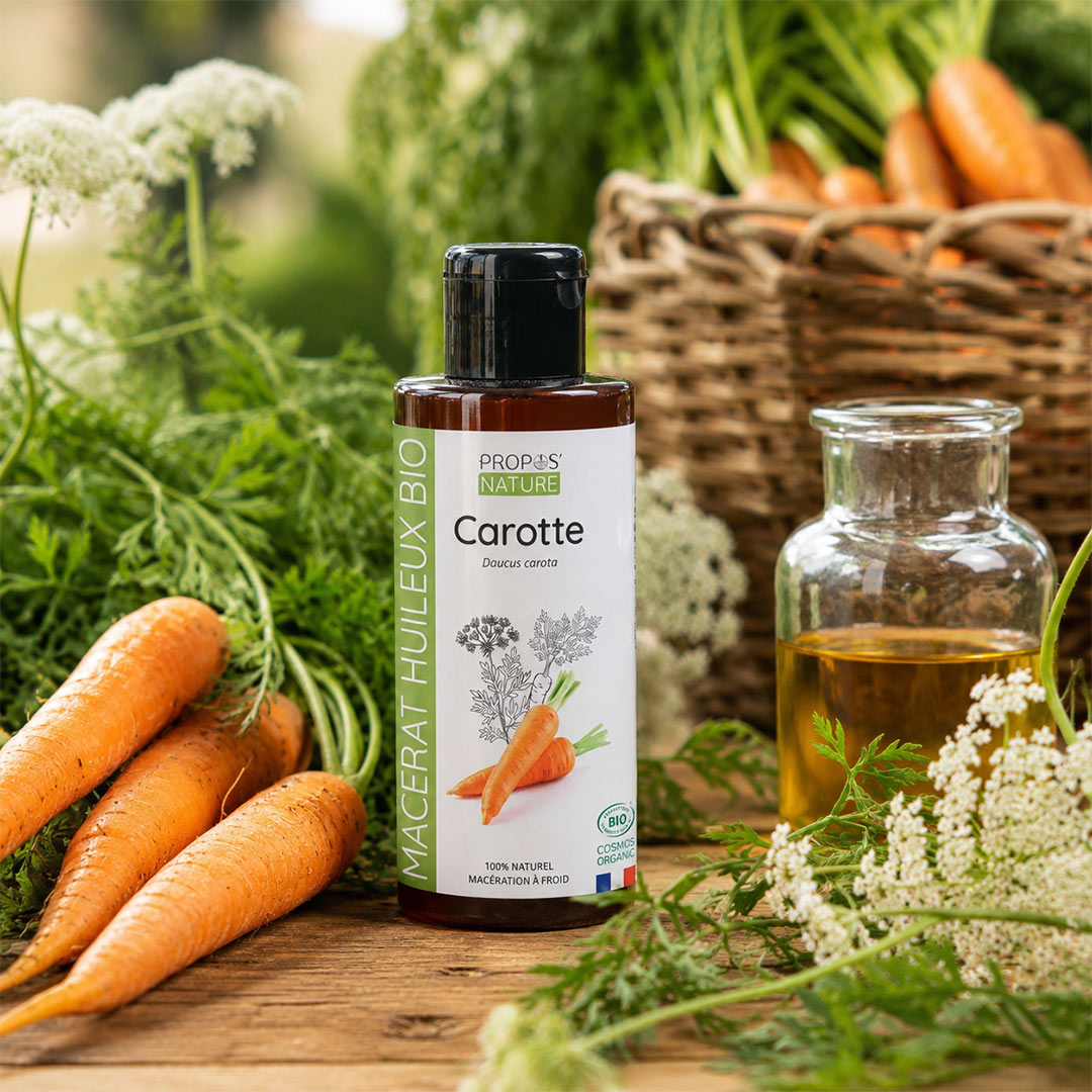 A bottle of PROPOS'S NATURE Carotte (carrot) organic macerate oil stands on a rustic wooden table surrounded by fresh carrots, green herbs, and white carrot flowers, with a glass bottle of the oil nearby.