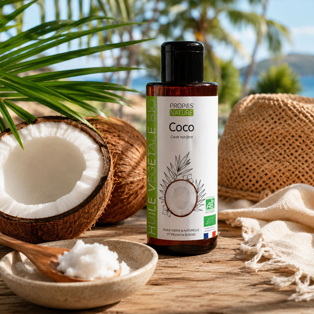 A bottle of Propos Nature Bio Coco Vegetable Oil stands on a wooden table surrounded by coconut halves, a wooden spoon with white coconut fat, a woven hat, and tropical palm leaves, with a blurred beach background.