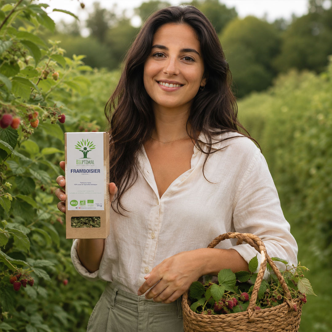 A woman standing in a raspberry field holds a box of Raspberry infusion and a woven basket filled with fresh berries and green leaves. The box has a green leaf logo and the brand name 'L'HEUREUSE'.