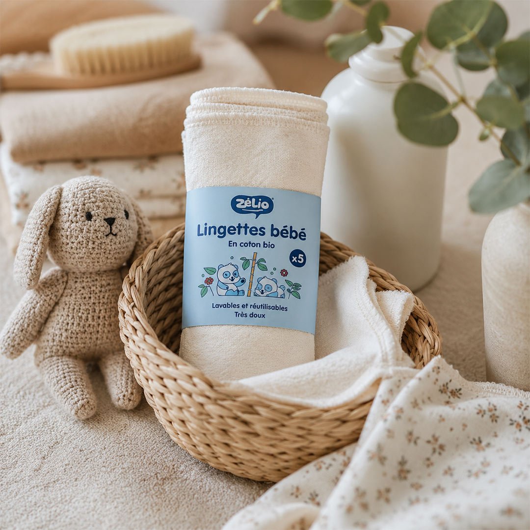 A set of soft baby washcloths in a woven basket, styled with a plush bunny toy, a wooden brush, and eucalyptus branches in a warm, neutral-toned setting. The brand 'Zélio' is visible on the label.