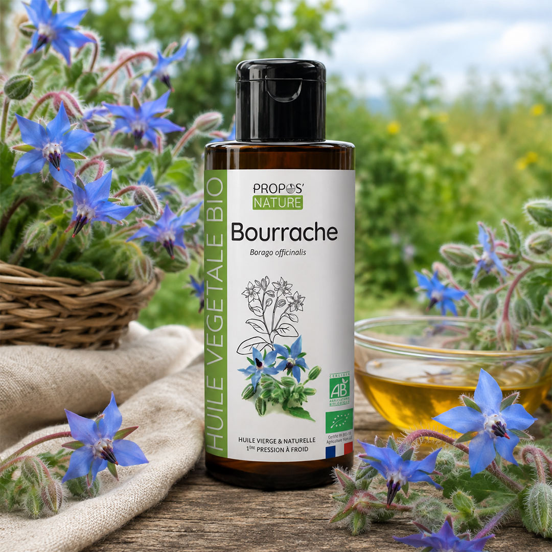 A bottle of Propos Nature Bio Bourrache oil stands on a rustic wooden surface outdoors, surrounded by blooming blue borage flowers, a wicker basket, a glass bowl of golden oil, and a soft blurred green garden background.