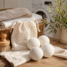 Four white wool dryer balls displayed on a wooden table next to a natural beige drawstring bag, a wicker basket with folded towels, a wooden brush, and a potted plant in a bright, airy laundry room setting.