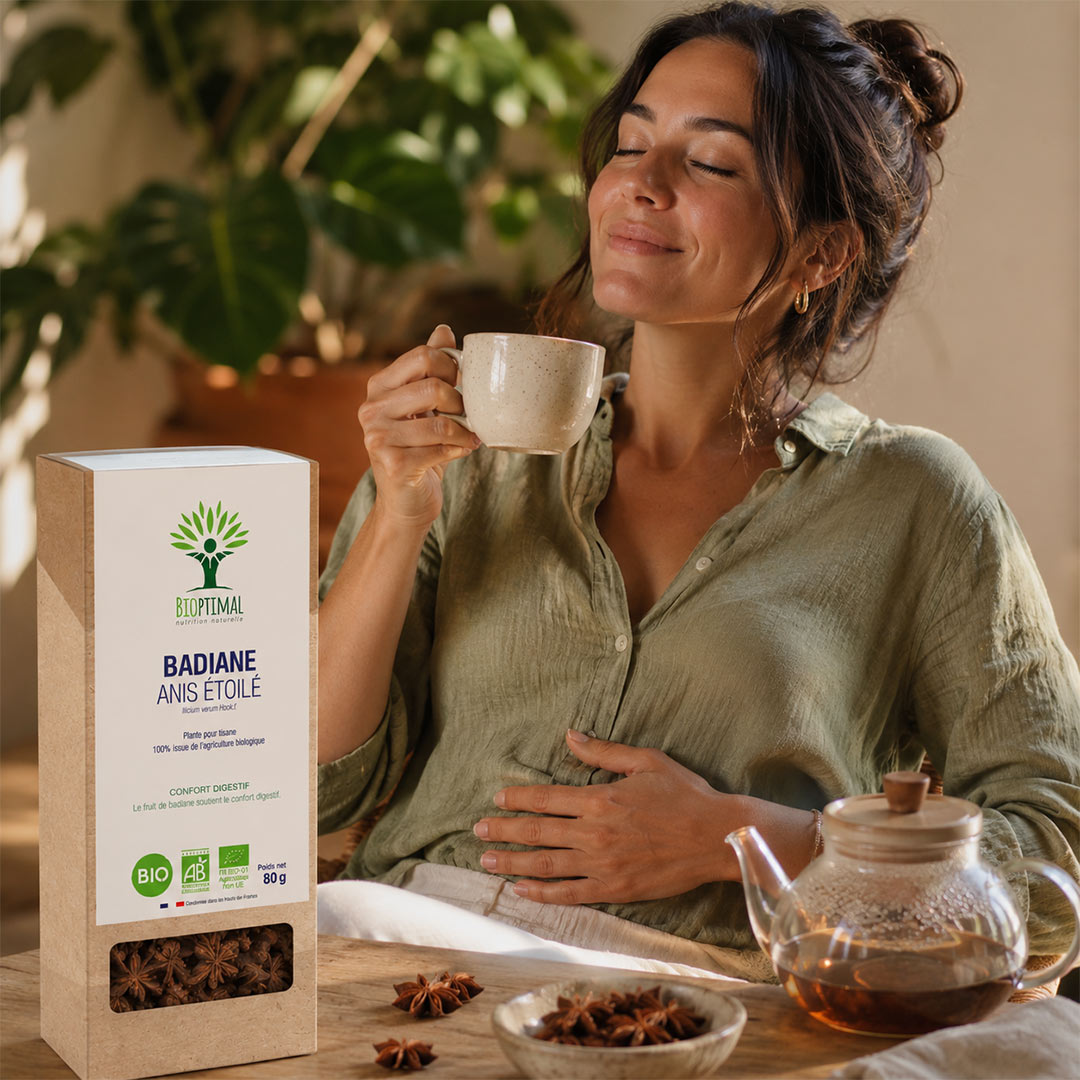 A woman in a sage green shirt holds a white mug with a pleasant expression, standing next to a box of BioPralmi Badiane Anis Etoile star anise tea. A glass teapot filled with tea and loose star anise sit on the wooden table in a warm, cozy setting with a blurred indoor garden background.
