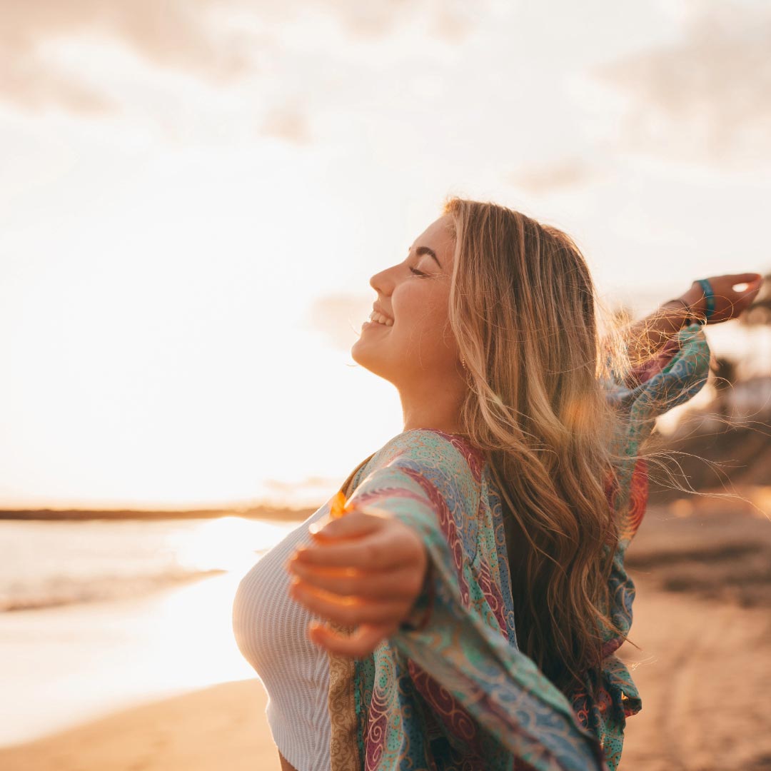 A woman smailing while holding her arms out, standing in sunset by the sea